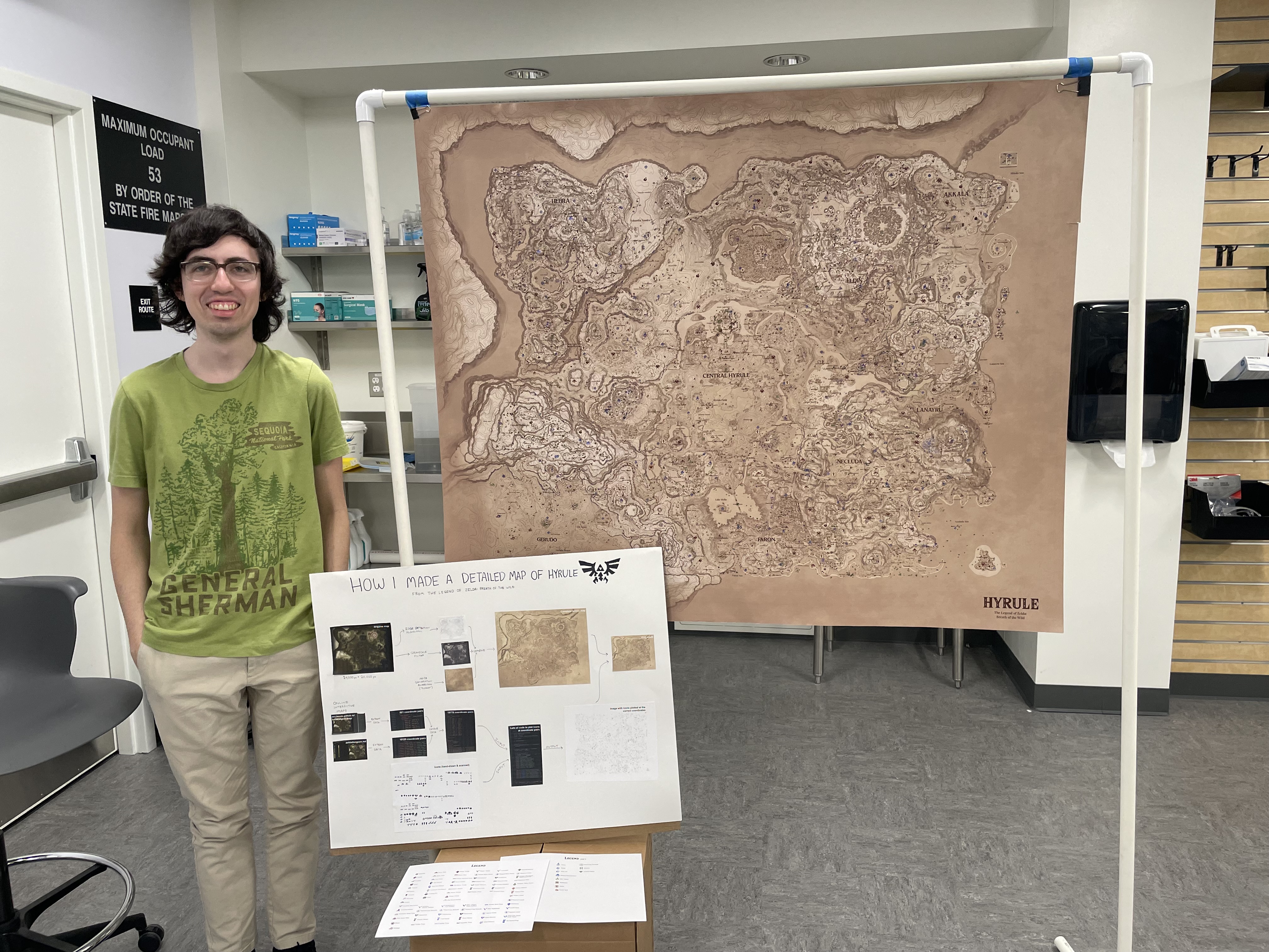 Me in a classroom standing next to my four-foot print of my map of Hyrule, suspended from a PVC pipe frame, and a flowchart showing the process of making the map